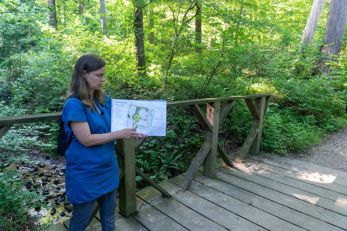An image of a woman pointing to a blueprint of Amazon's HQ2 while standing on a bridge on a hiking trail with green trees surrounding her.