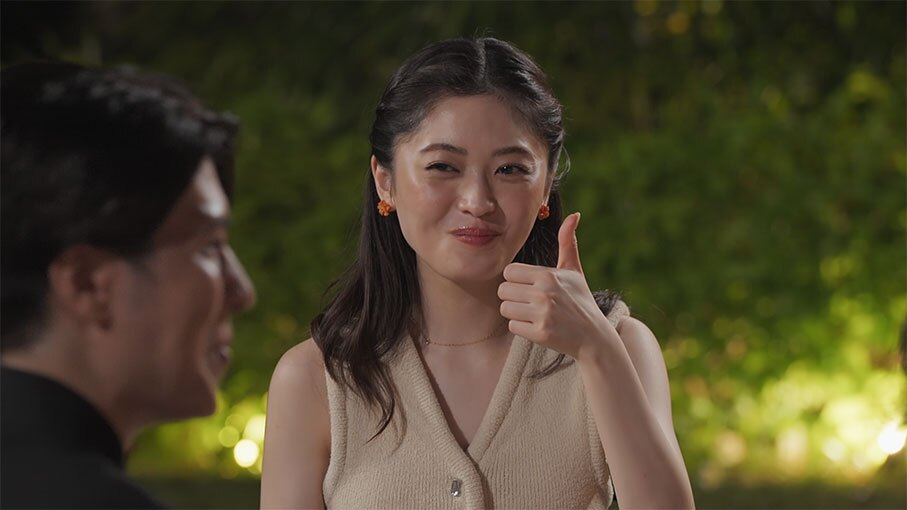 薄暗い屋外、ライトアップされた植栽を背景に笑顔で親指を立てる女性。女性の視線の先、左側に男性の横顔。