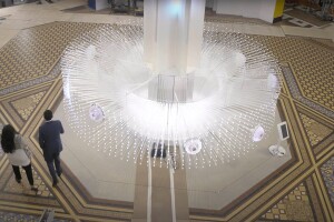 An image of a large white structure with multiple glass shoots coming out of it. At the end of each shoot is a small light. There are small circles within the structure that have smaller clusters of lights with a purple tint. The structure stands in the middle of a museum, and two people are walking by it.