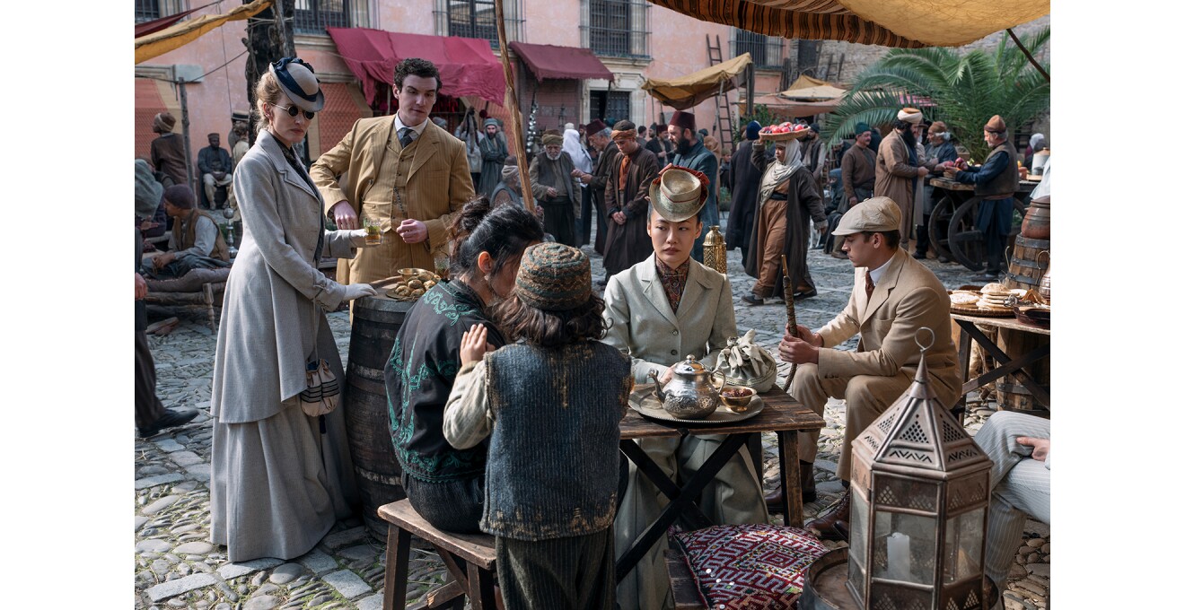 中東風のマーケットのような屋外で、テーブルに向って腰掛けた女性。手前に座る2人の人物を見つめている。右側にも男性が1人座り、左側には男性と女性が1人ずつ立っている。