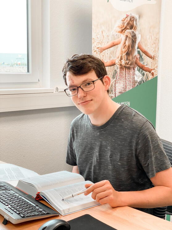 Ein Mann mit grauem T-shirt sitzt vor einem Computer und lächelt in die Kamera.