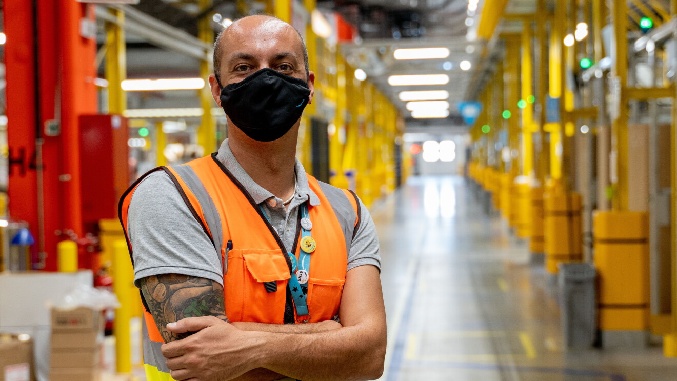 Oreste Tagliaferri, director general de los centros de distribución de España e Italia. En un pasillo del centro logístico está con los brazos cruzados Oreste. El pasillo tiene estanterías metálicas de color amarillo. Oreste tiene el brazo derecho tatuado. tiene el pelo muy corto y la parte superior no tiene pelo. Lleva una mascarilla de Amazon de color negro, un chaleco de color naranja y un polo de color gris. 