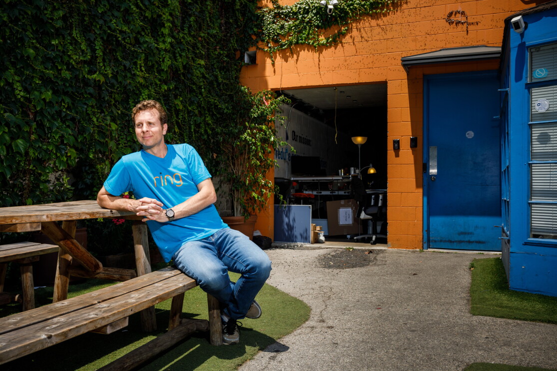 Jamie Siminoff, founder of Ring, sits at a picnic table at Ring headquarters in Santa Monica, CA. He is in a blue t-shirt and jeans, and is looking to the left. 
