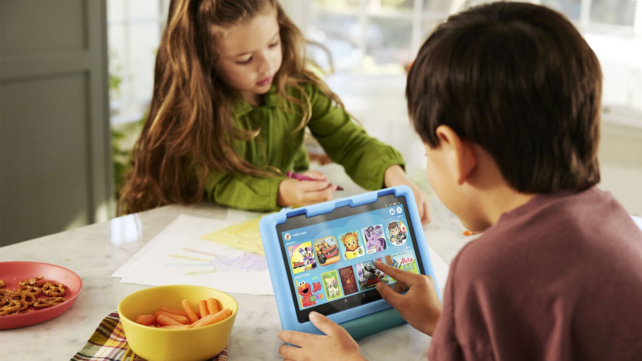 An image of a boy and a girl sitting on the table. The girl is drawing with a crayon and the boy is interacting with a tablet, which shows Amazon Kids on the screen.