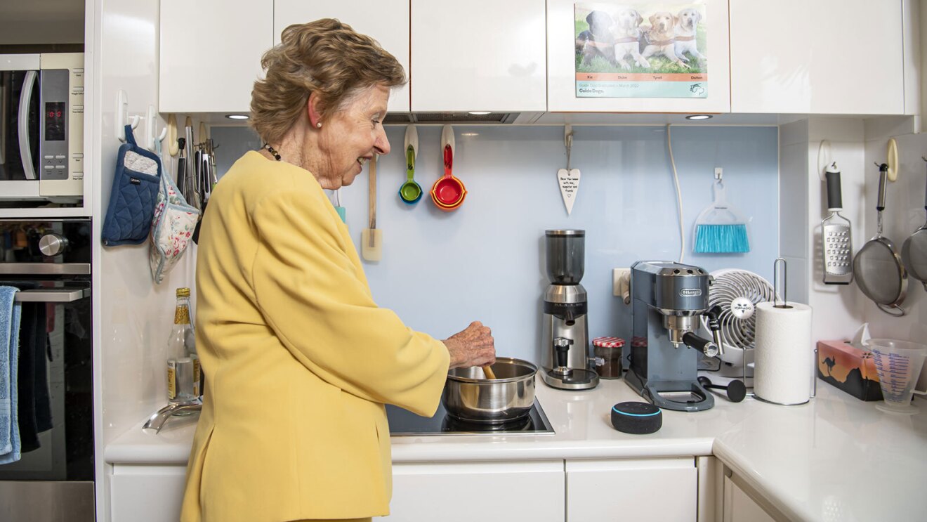 An image of a woman wearing a yellow shirt cooking something in her kitchen with an Echo device next to her.