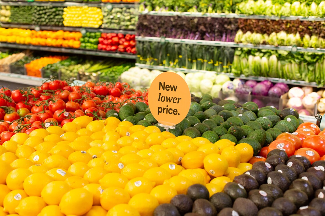 Inside a Whole Foods Market store, lemons, limes, avocados, and tomatoes are stacked on a table, with a "New lower price" sign in the center of the produce. Behind the display, lettuces, peppers, squash and other vegetables are stocked in a produce wall display. 