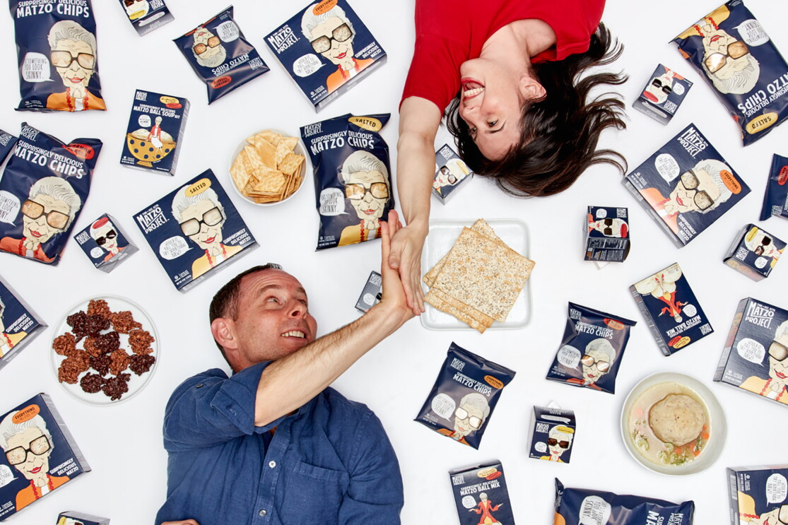 A woman lies on her back, with her right hand extended above her head as she gives a man, also lying on the ground, a high five. Around them are packages of matzo and other products.