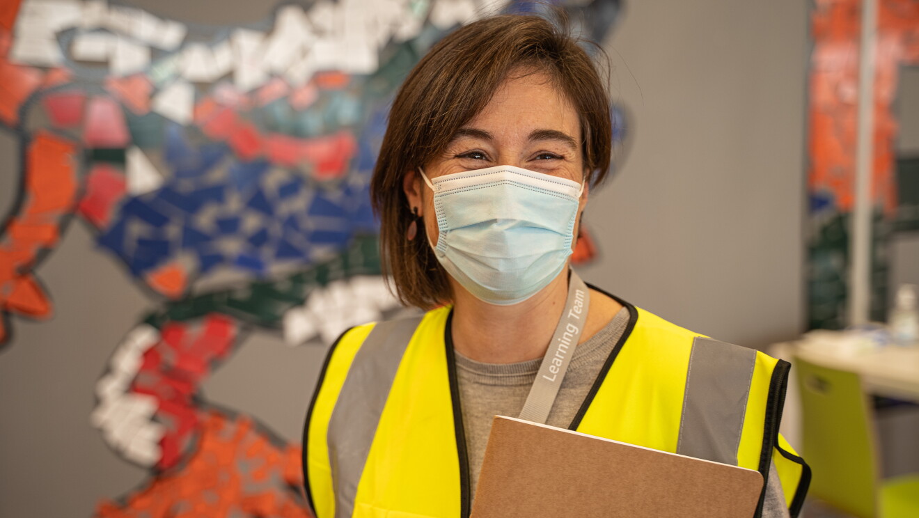 Marta Casasayas, responsable del programa de Aprendizaje de Amazon en España. De fondo una pare de colores. Marta sonriente y con mascarilla mira a c´ámara, sujeranto con la mano izquierda una carpeta de Amazon. Tiene media melena castaña pero de un lado es más corta que del otro. 