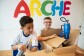 Zwei Kinder packen Pakete aus und stehen vor der Aufschrift "Arche". 