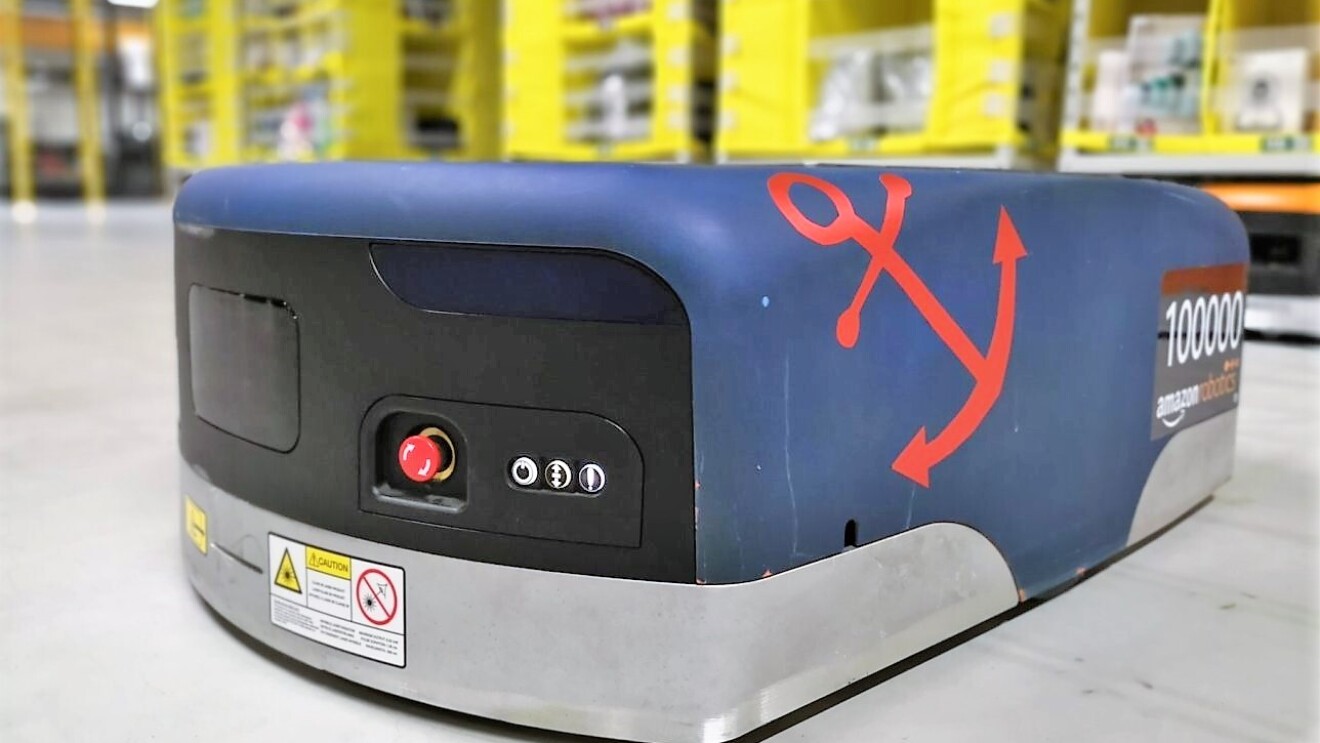 Auf dem Boden einer Lagerhalle sieht man einen blauen Amazon Transportroboter. Auf der rechten Seite wurde ein roter Anker aufgeklebt und ebenfalls rechts steht die kennzeichnung "100000 amazon robotics".
