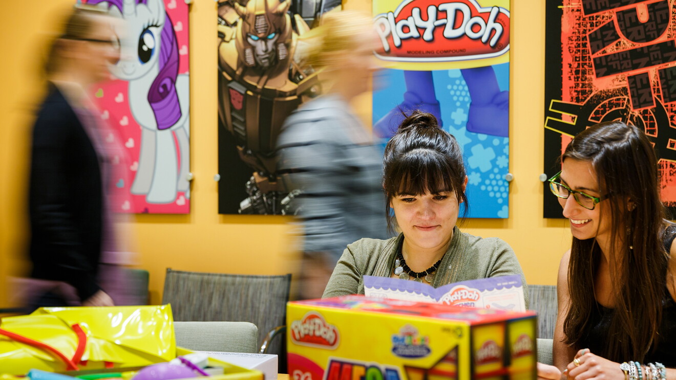 Deux femmes au premier plan sont assises à une table entourée d'emballages. D'autres personnes passent en arrière-plan. Le mur contient des affiches de My Little Pony, Play-Doh et d’autres marques Hasbro.