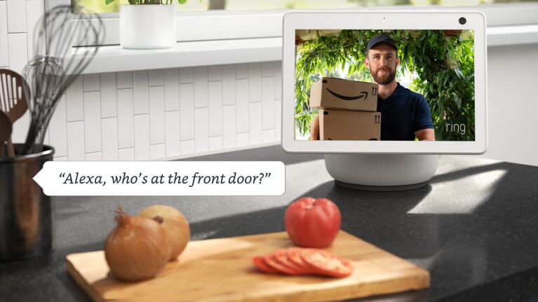 An image of an Echo show device sitting on a kitchen counter next to a cutting table with assorted vegetables. The screen is showing a person delivering Amazon packages through the Ring camera, and there is a text box on the left that reads "Alexa, who's at the front door?"
