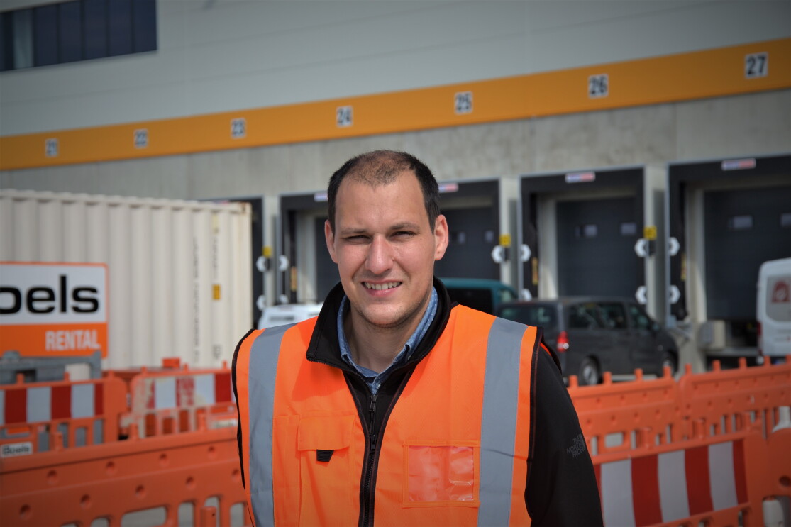 Ein Mann mit braunen kurzen Haaren und orangener Sicherheitsweste vor neinem Logistikzentrum. Man sieht Absperrungen und die Laderampen.