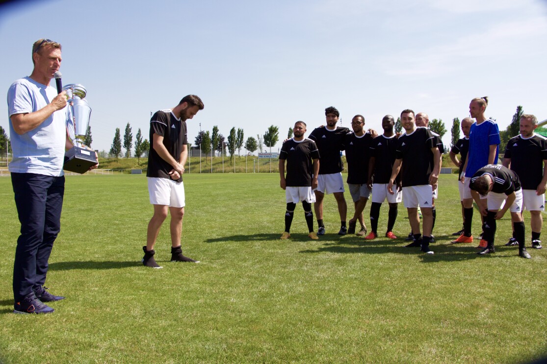Ein Mann rechts im Bild hält einen Pokal. Um ihn herum versammeln sich Fußballspieler.