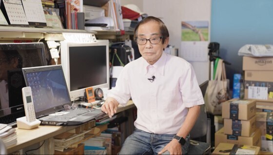 事務所兼倉庫のような室内で、椅子に座る男性。デスクにはノートパソコンやモニターがおこあれている。