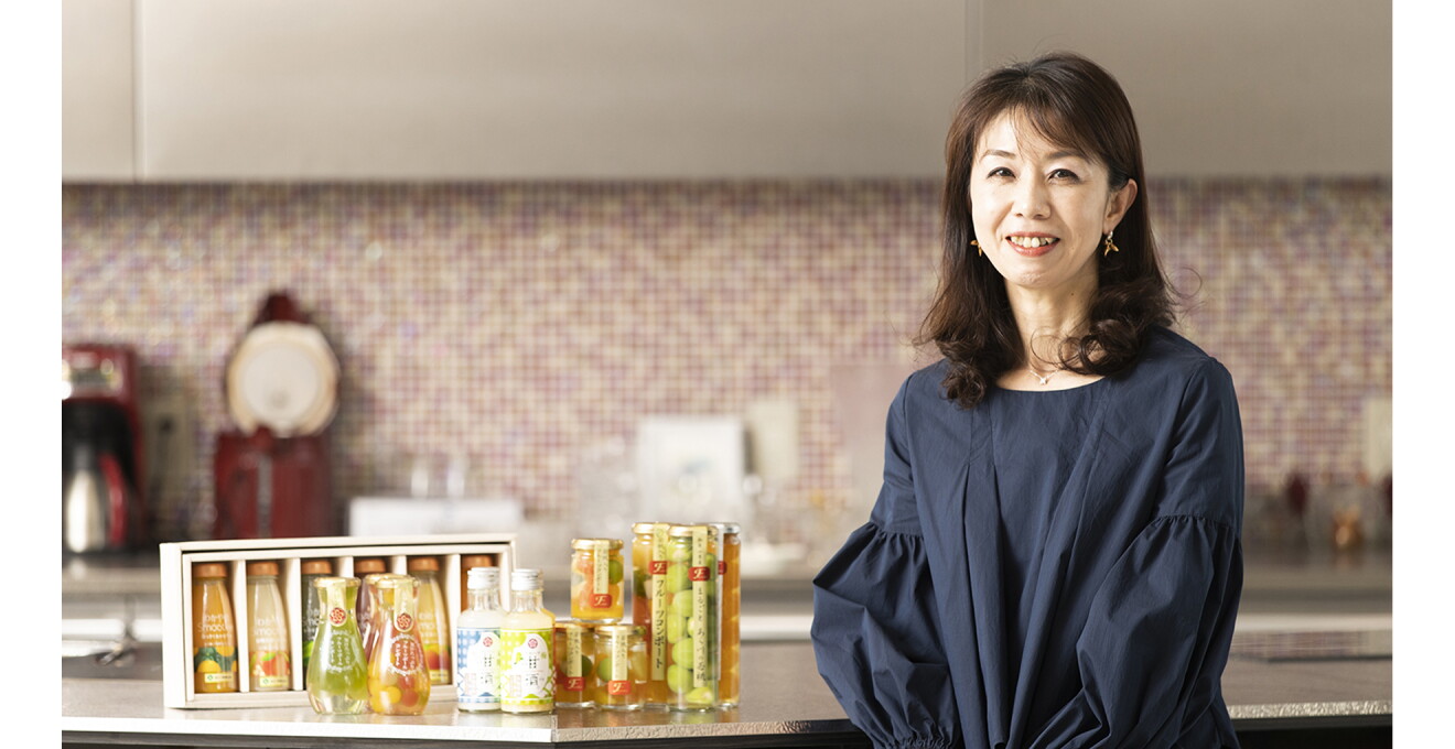 株式会社ふみこ農園 専務取締役の成戸晃子さんと、スムージー、甘酒、フルーツコンポートなどの商品がならんだ写真