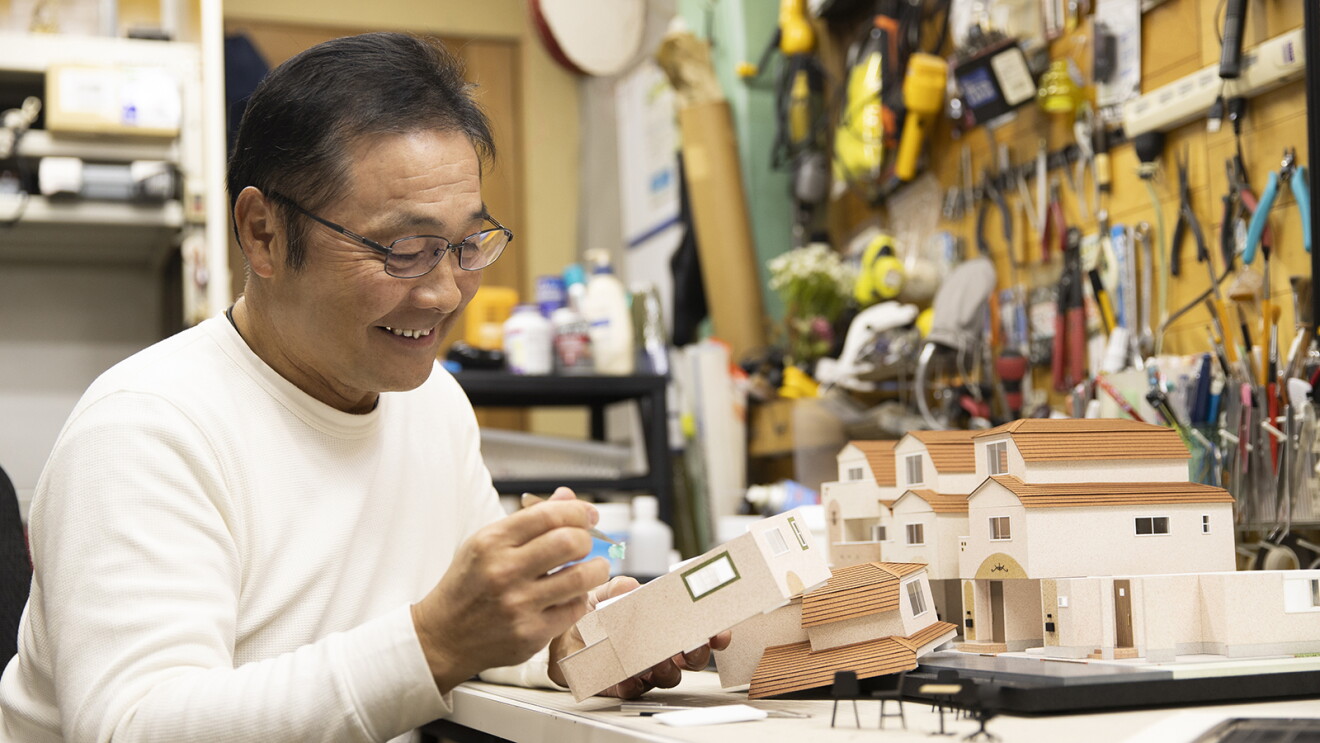 さまざまな道具がかかる壁の前で住宅の模型を組み立てている男性