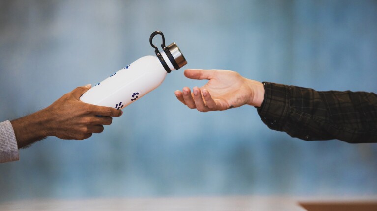 A man wearing a gray waffle weave shirt hands a white water bottle across a counter, to a man wearing a plaid button down.