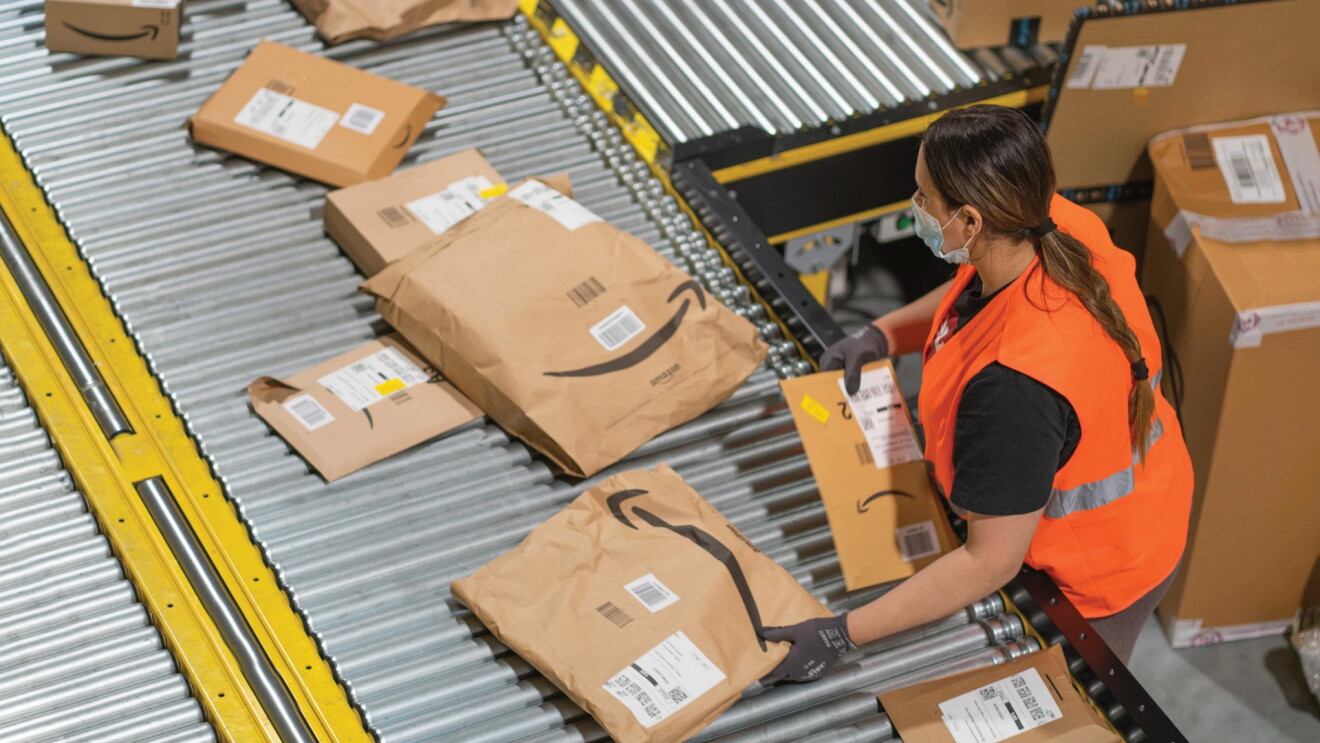 A woman wearing a safety vest stands to the right of a conveyor belt. She is handling flat packages, moving them along the belt.