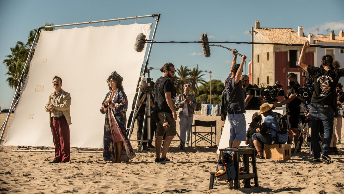 En un set de rodaje exterior, en la arena de la playa, aparecen dos actores, hombre y mujer vestidos de los años 60. Una pantalla blanca para aportiguar la luz. De fondo dos casa de pueblo marinero y, al lado de los actores, un equipo técnico de unas 10 personas con cámaras y micros. Todos van vestidos con ropa oscura.