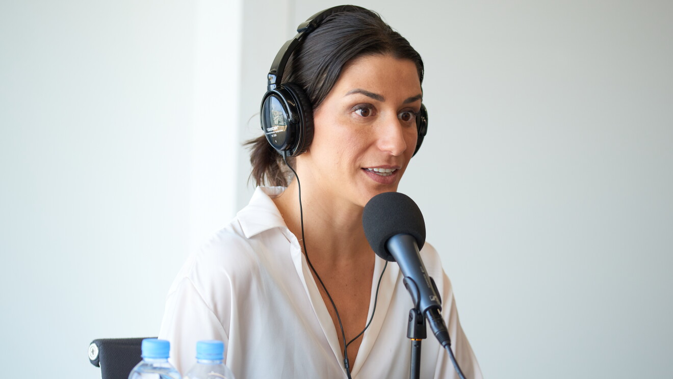 Amanda Machado, CEO de Andrés Machado, sentada en una mesa delante de un micrófono y con unos auriculares puestos. Ella está mirando al infinito. Lleva el pelo largo con pelo castaño. Con una camisa  estampada con puntos y cuadrados dibujados y pinceladas de colors como morado, verde, rosa o naranja.