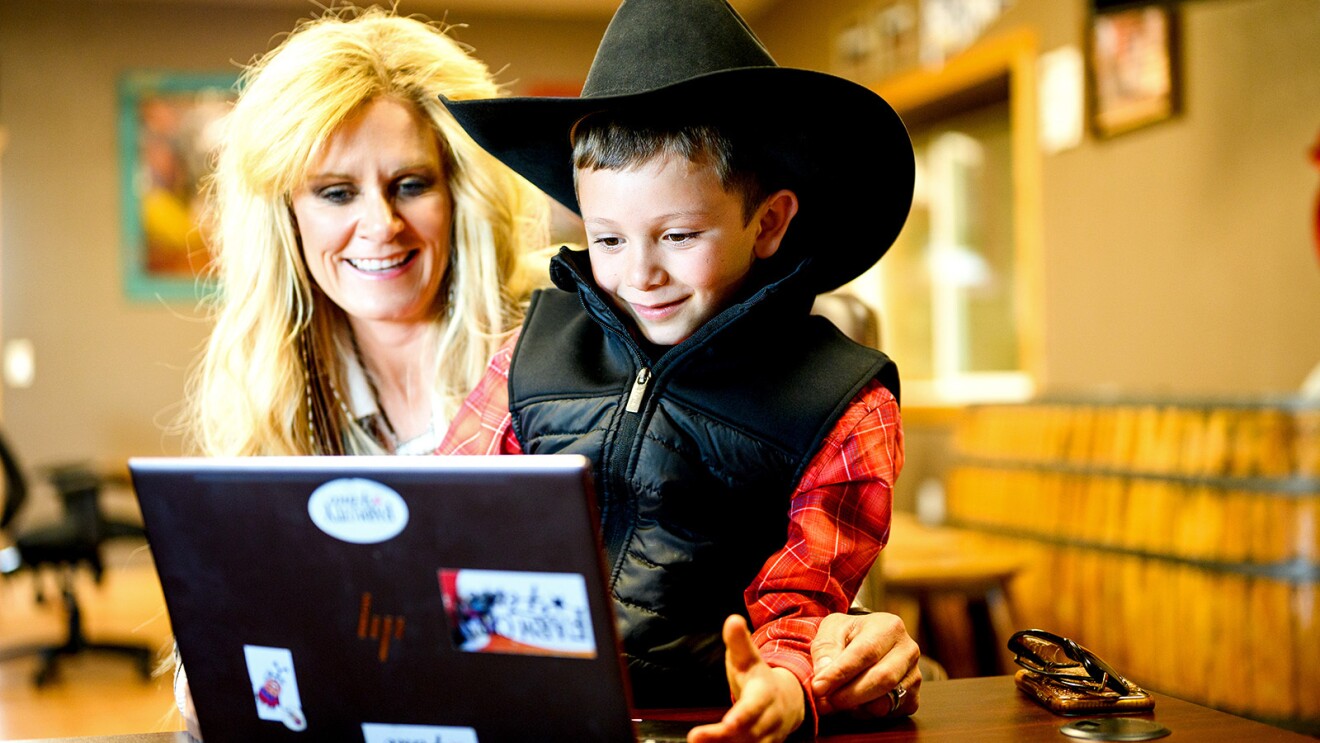 A photo of a mother and her son sitting at a desk looking at a laptop device.