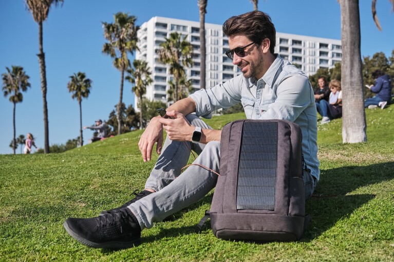 Ein Mann sitzt mit seinem Handy und einem Sunnybag, einem klimaschonend hergestellten Rucksack mit integrierten Solarpaneelen, auf einer grünen Wiese in der Sonne.