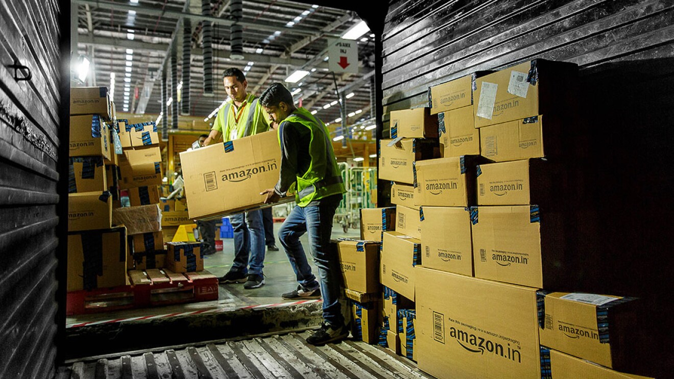 Two Amazon associates are working to load a truck with packages to be delivered to customers. They are both holding onto a large box as one of the associates steps into the trailer of a truck. Within the truck, there is a wall of packages, prepared for delivery.