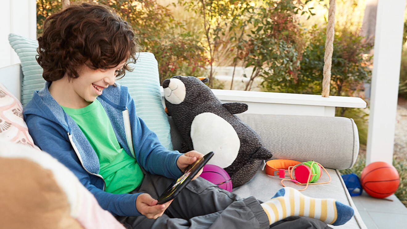 一个男孩坐在院子里的摇椅上,穿着绿色衬衫和蓝色毛衣,看着他手里的平板电脑。他旁边有毛绒玩具、球和耳机。