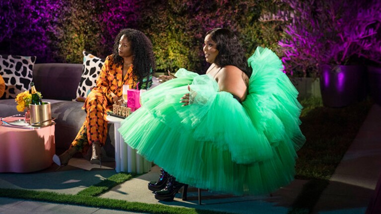 An image of singer Lizzo wearing a green dress and having a sit-down conversation. Next to her is a woman wearing an orange jumpsuit. There are trees and pink lights in the background.