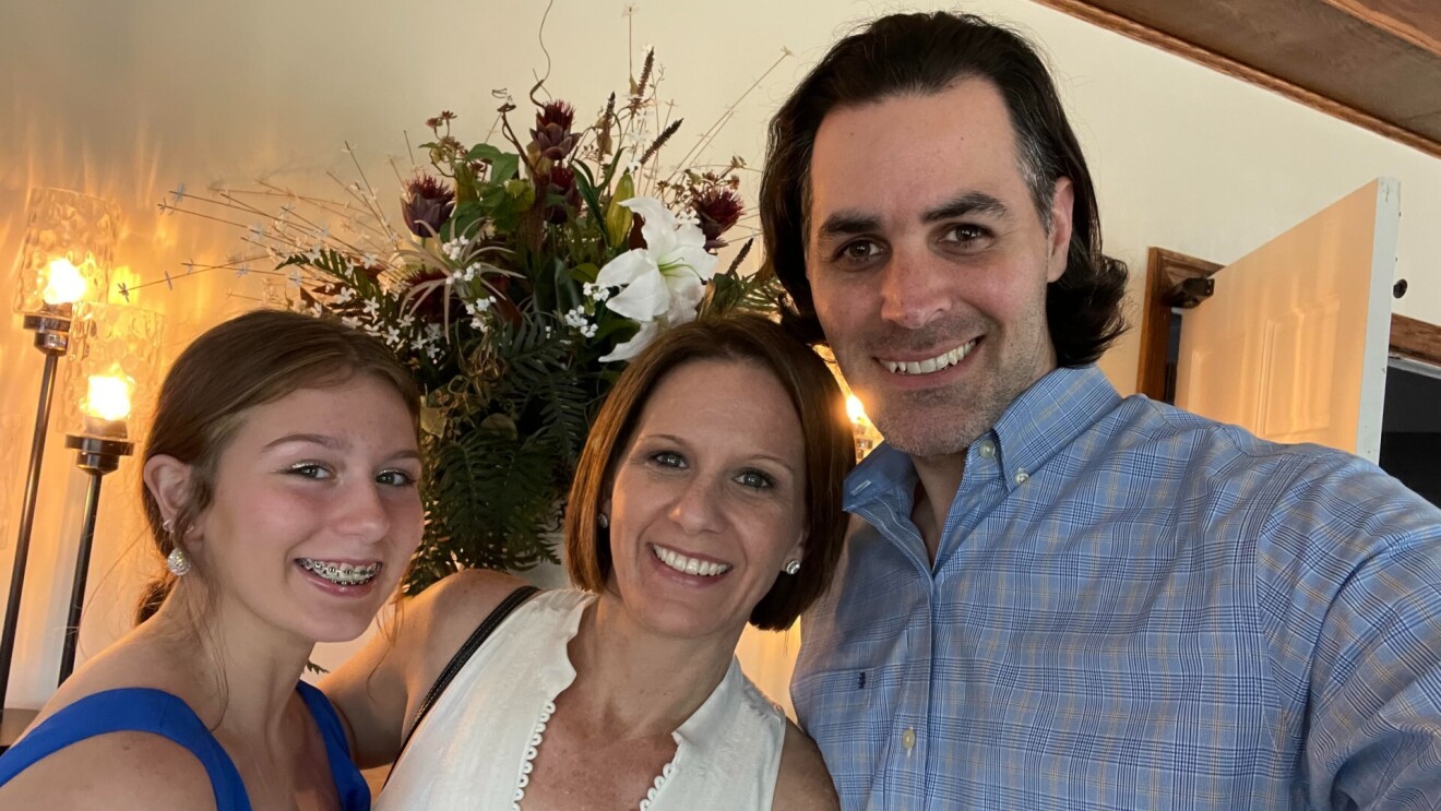 A father, mother, and daughter smile with their arms around one another.