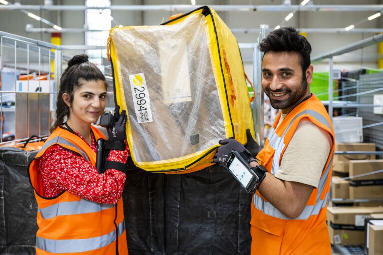 Zwei Amazon Mitarbeiter:innen (eine weibliche Mitarbeiter und eine männliche) halten einen gelben Transportbehälter in den Händen.