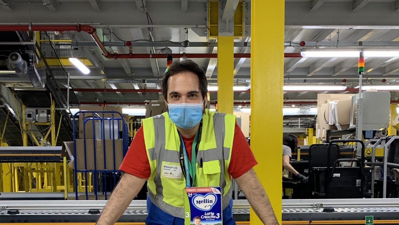 Volontario Amazon sorride dietro la mascherina all'interno di un centro di distribuzione. E' poggiato con le braccia su una grossa scatola indirizzata alla Croce Rossa Italiana.