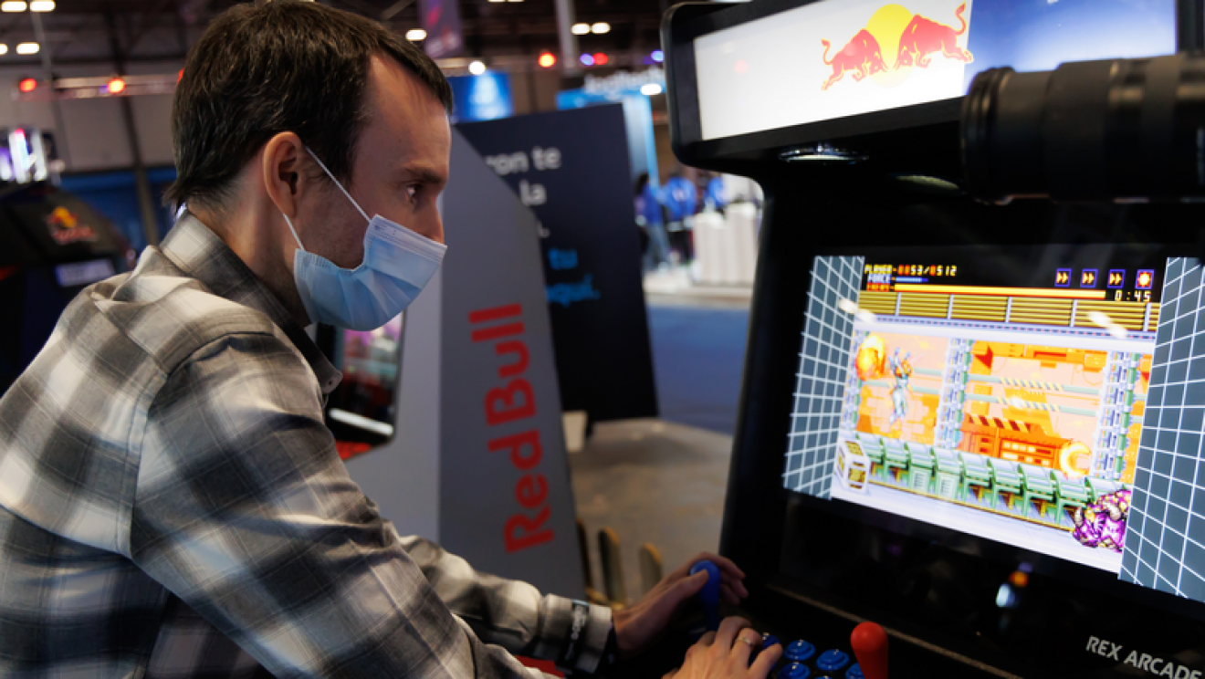 Eduardo García, responsable de Gaming y esports de Amazon Retail Europa. Eduardo con el pelo corto negro y mascarilla quirúrgica jugando a videjojuegos. Lleva una camisa de cuadros grises, negros y blancos. 