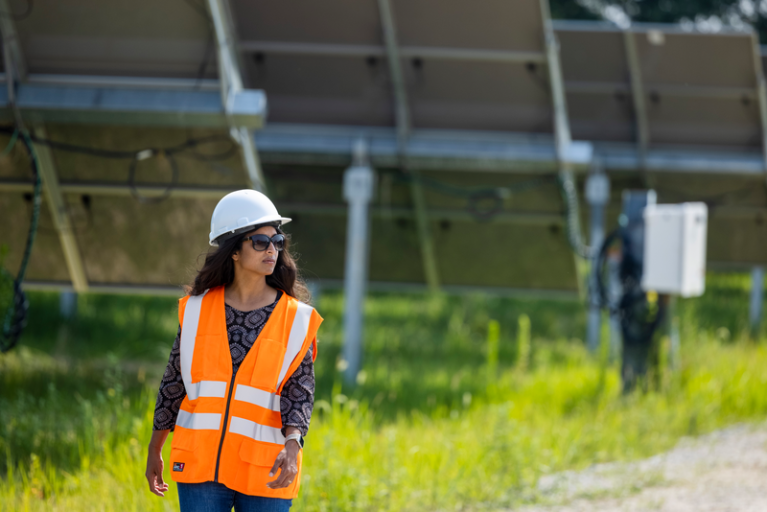 En una granja solar, delante de la parte trasera de tres placas aparece caminando una mujer con gafas de sol, casco blanco, y el pelo ondulado largo. Lleva una jeans, un chaleco de color naranja con un jersey negro estampados con formas geométricas grises.