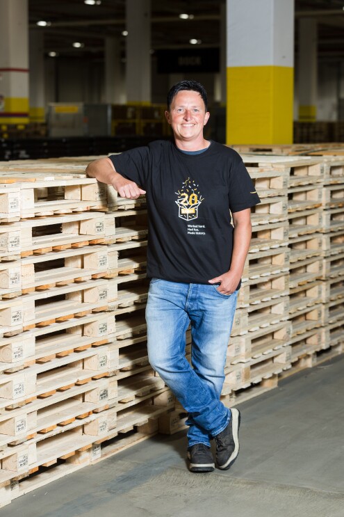 Eine Frau mit dunklem Kurzhaarschnitt und einem schwarzen T-Shirt, auf dem "20 Jahre" zu lesen ist, lehnt lächelnd an gestapelten Paletten.