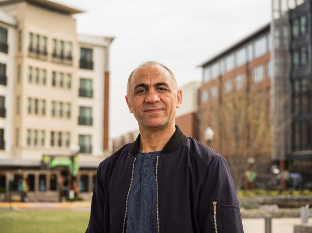 Khalid Noorzad stands outside in a park between buildings and grins at the camera.