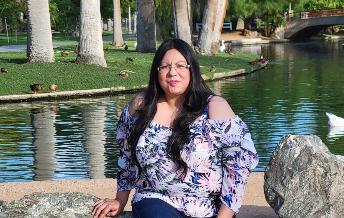 An image of a woman smiling for a photo while sitting on a bench in front of a pond at a park.