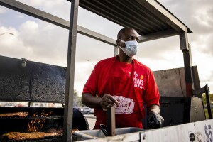 A man stands in front of an outdoor smoker. He wears a mask and holds products to prepare meals for customers. 