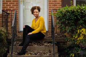 A woman, Crystal Swain-Bates, sits on the front step of a brick home. She waers black booties, black pants, a mustard yellow sweater, and large good hoops. 