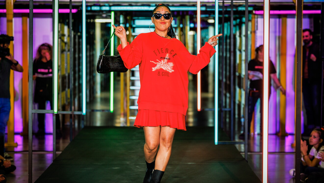Dentro de un centro logístico, con las estanterías de fondo y las barandillas de color amarillo, aparece una pasarela de color negro ambientada con luces de neón y fotos de trabajadores de Amazon posando. Lilimel Rivas lleva un vestido rojo, botas militares, bolso de mano y gafas cat eye. Lleva la cara maquillada con pinceladas de los colores de la bandera de su país. En los extremos de la pasarela hay niños con camisetas blancas de manga corta mirando a sus padres desfilar. 