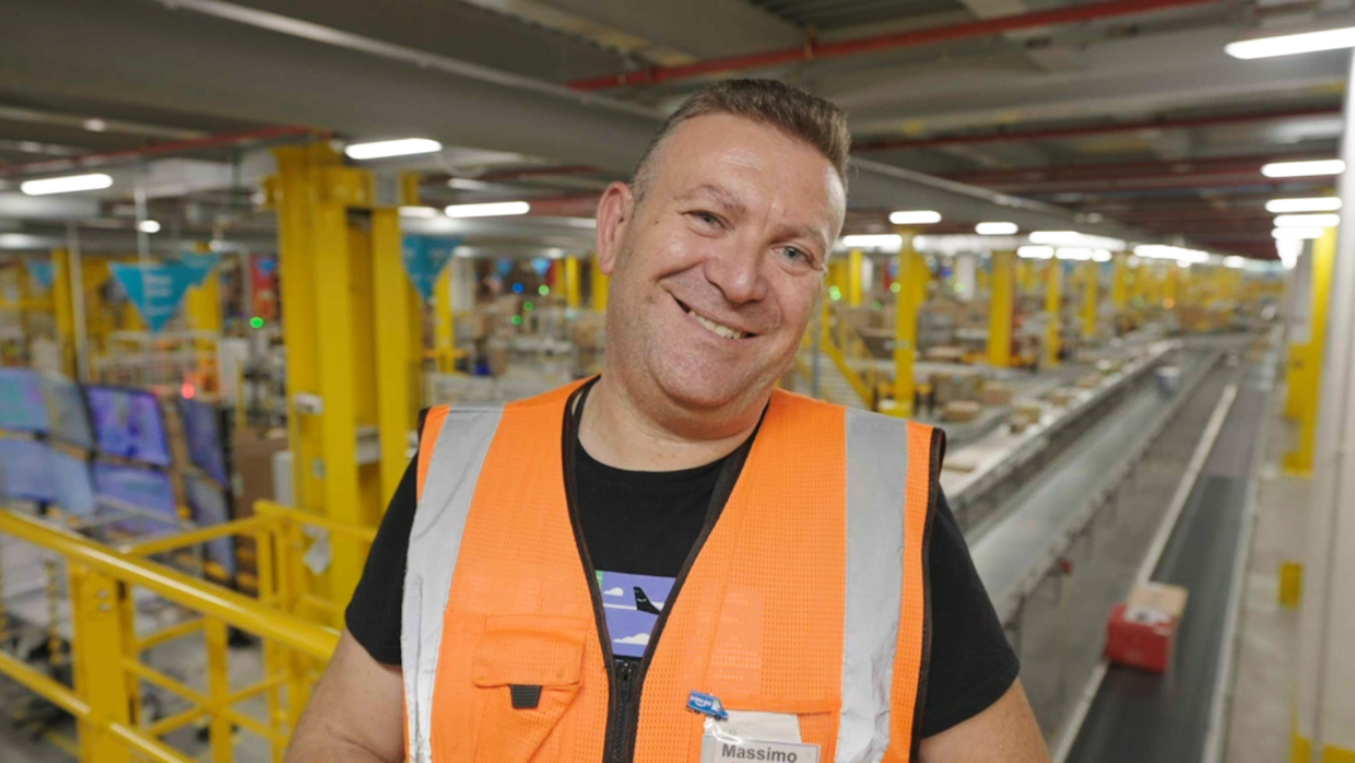 Primo piano di un dipendente Amazon all'interno di un magazzin. L'uomo sorride, ha i capelli corti e indossa una t-shirt nera sotto un giubbino di sicurezza arancione