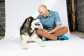 A man and a dog sit together, making eye contact, on a white photography background.