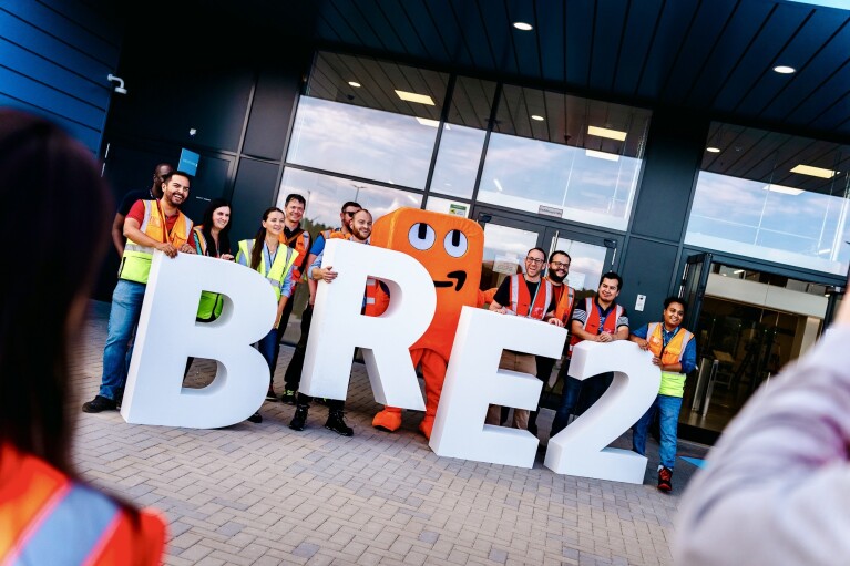 Amazon Mitarbeiter:innen stehen mit dem orangenen Amazon Maskottchen "Peccy" hinter weißen Großbuchstaben BRE2. Das Kürzel steht für das Logistikzentrum Großenkneten und BRE2 für den nächstgelegenen Flughafen in Bremen.
