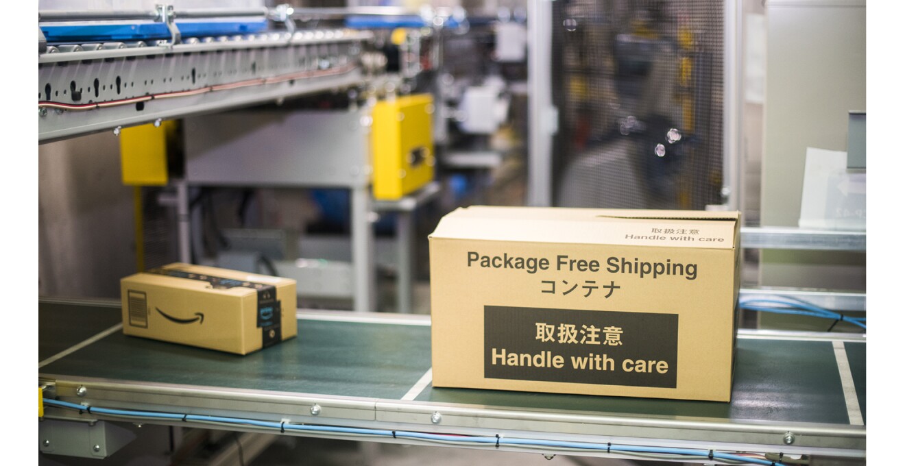 FC内のベルトコンベヤーに「Package Free Shipping・取扱注意」と書かれた簡易包装商品を保護するコンテナが流れている