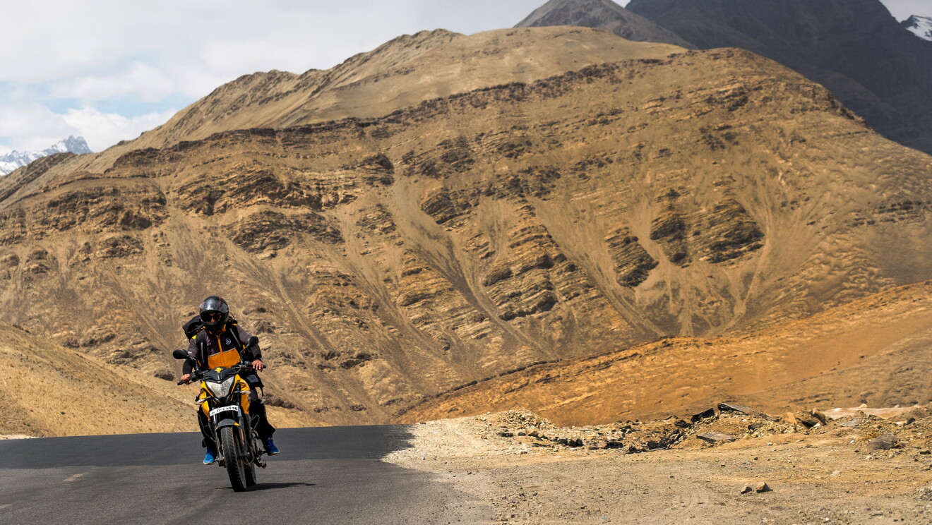 A motorcyclist rides on an asphalt road. Behind him, the road curves to the right and disappears from view. In the background a tan rock formation rises up from the road.