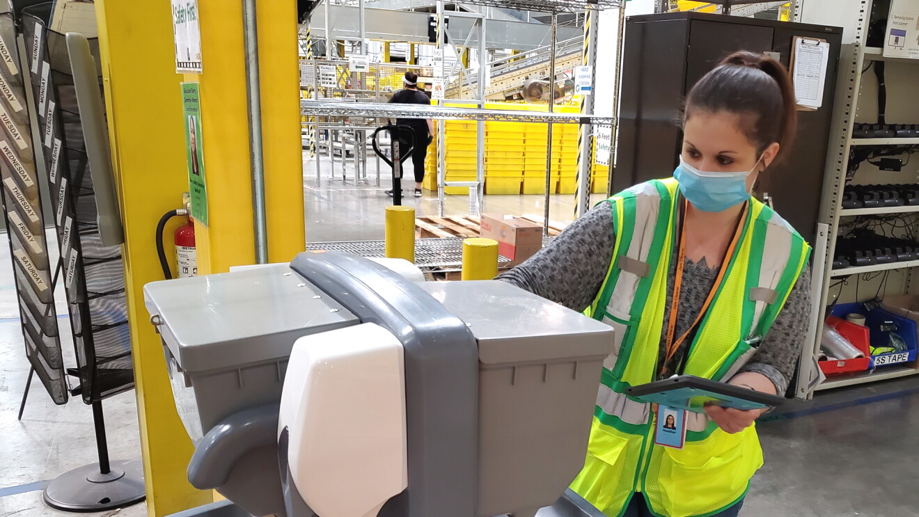 A woman wearing a safety vest and mask inspects a hand-washing station.