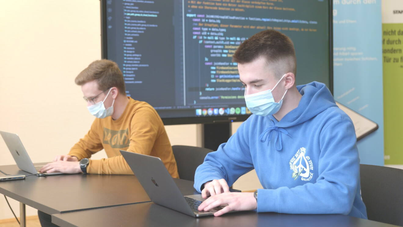 Zwei Jungen mit Masken sitzen an einem Tisch und schauen in ihre Laptops. Im Hintergrund ist eine Leinwand mit Computerbildschirm sichtbar.