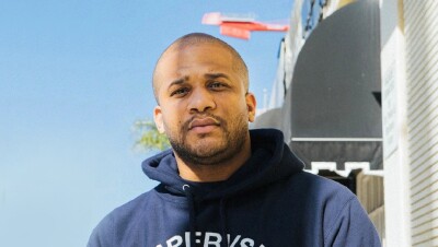 一个男人站在外面拍照的画面。在他身后有一棵棕榈树和一幅街景。