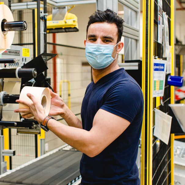 Ein junger Mann mit Maske an einer Etikettiermaschine
